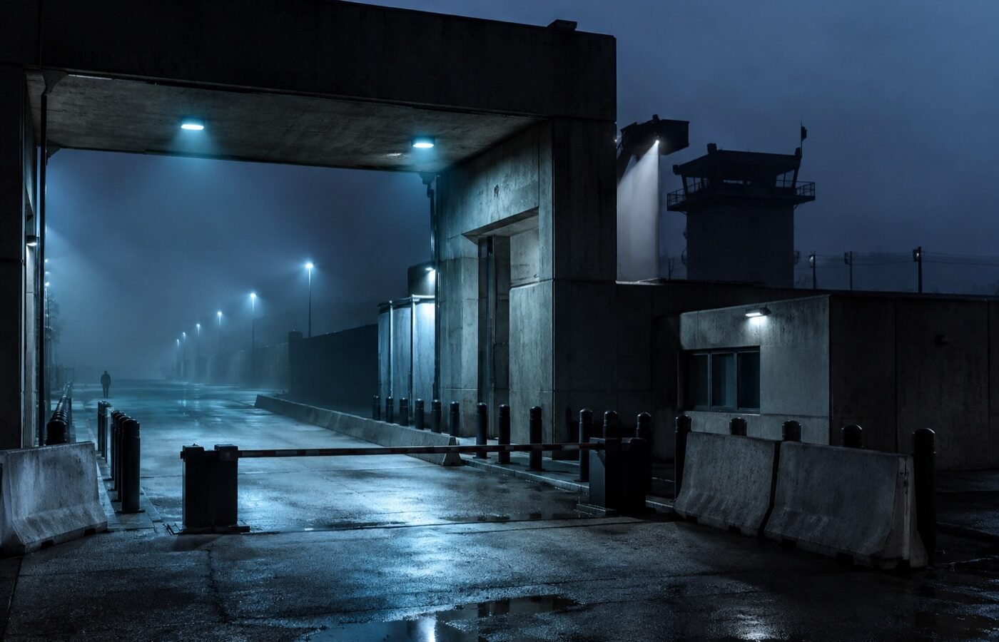 Hardened facility checkpoint with blast barriers and boom gate at dusk